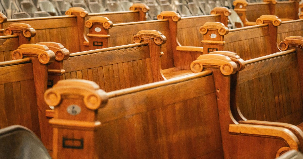 The image depicts several rows of wooden pews, commonly found in a church or a similar setting. The pews are made of polished, rich-brown wood with ornate details on the armrests, featuring circular carvings and small plaques. The seats have vertically aligned wooden slats along the backrests. The overall arrangement suggests a formal seating arrangement, and the perspective highlights a pattern created by the repeating shapes of the pews. The background shows additional rows of pews, partially blurred.